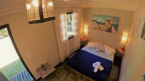A bedroom with a bed, a small table, a nightstand, a painting, and a window with curtains. - Morada do Sol Guest House (Búzios)