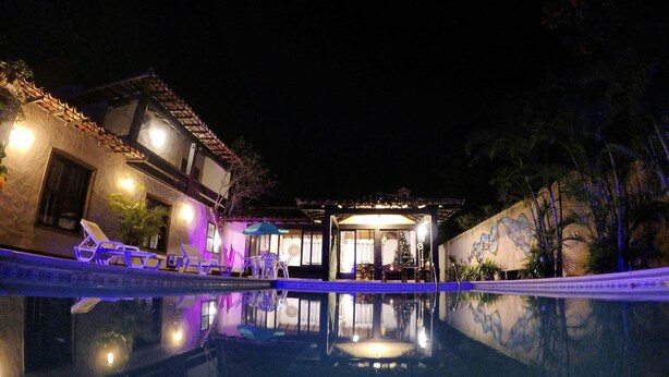 A nighttime view of a hotel with a swimming pool, lounge chairs, and a covered patio area. - Morada do Sol Guest House (Búzios)