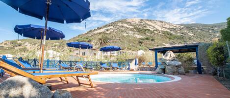 A pool area with lounge chairs and umbrellas, a stone structure, and a mountainous backdrop.
