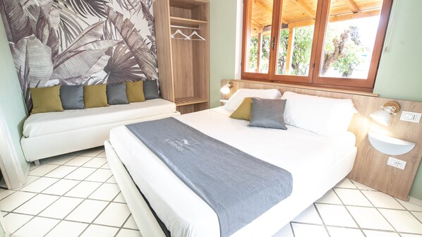 A bedroom with a bed, a sofa, a wardrobe, and a window with a view of trees. - Hotel Il Perseo (Marciana)