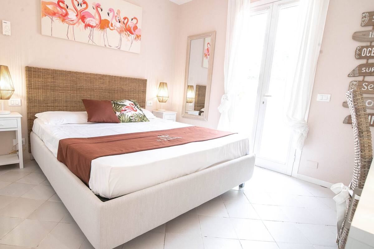 A spacious bedroom with a large bed, two bedside tables, a wall-mounted mirror, and a beach-themed sign on the wall.
