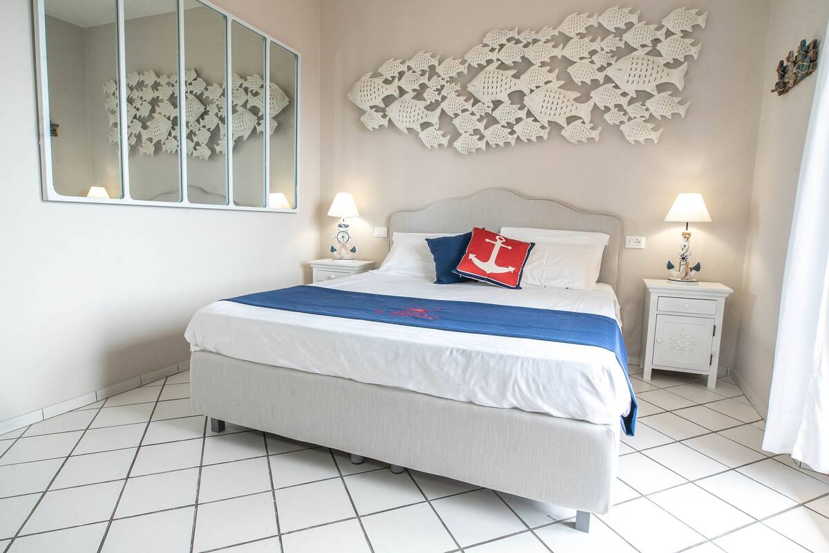 A neatly arranged bedroom with a large bed, two bedside tables, a wall-mounted mirror, and a decorative fish wall art piece.