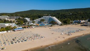 A beach with numerous umbrellas and sun loungers, a large hotel building, and a forested hill in the background. - MARITIM Paradise Blue Hotel & Spa (Albena)