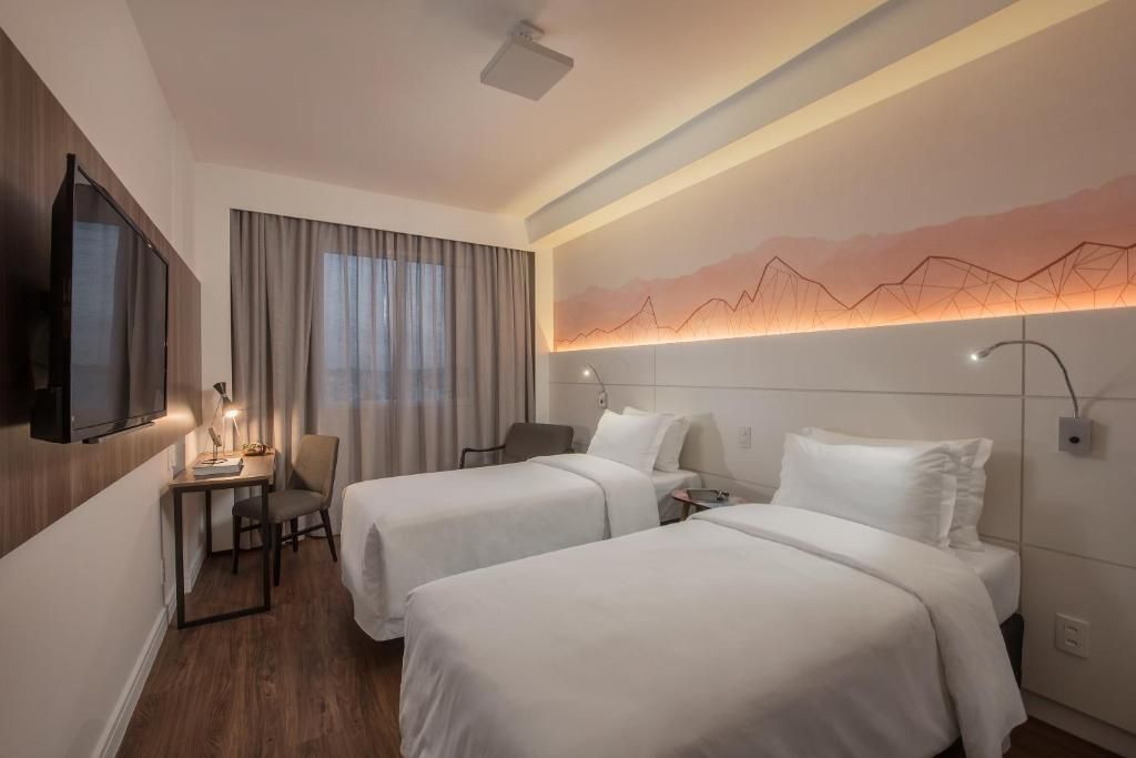 Habitación de hotel con dos camas, un escritorio, una silla, un televisor y una lámpara fijada a la pared.