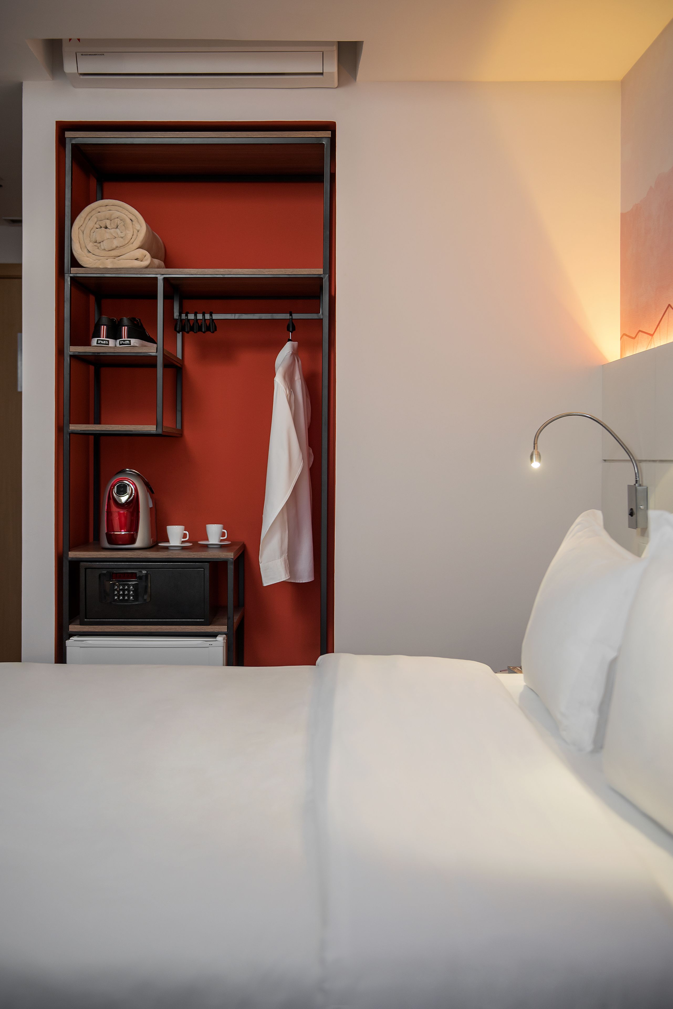 Een hotelkamer met een bed, een rode kast met een waterkoker, bekers en een handdoek, en een wandlamp.