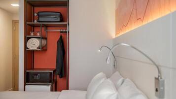 Habitación de hotel con cama, armario con abrigo colgado, microondas y lámpara fijada a la pared.