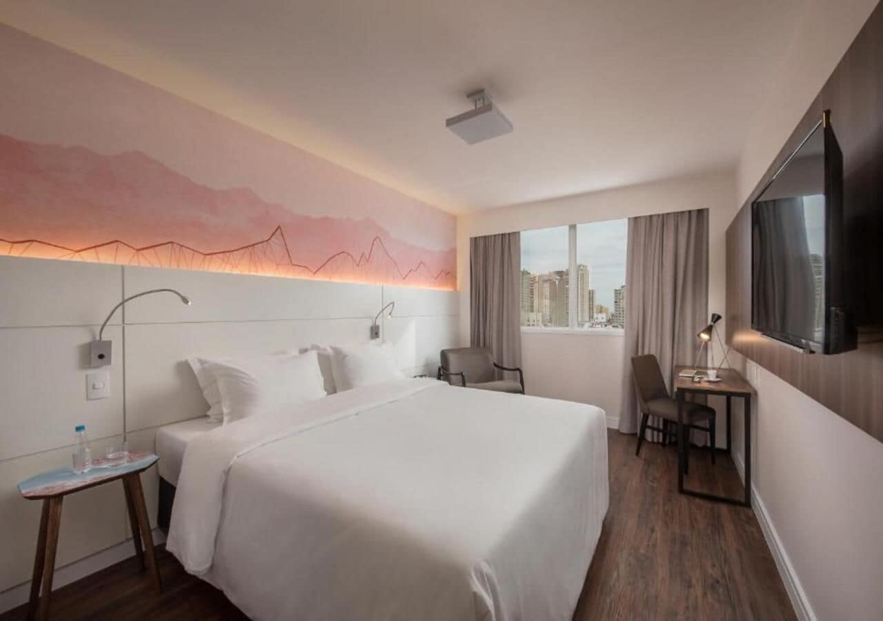 Habitación de hotel con cama, escritorio, silla, televisión y vista a la ciudad.