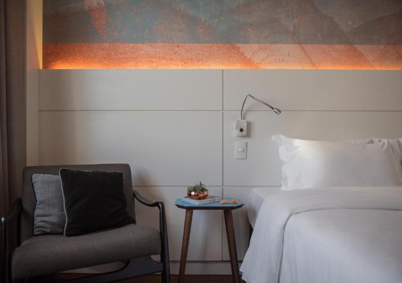 A hotel room with a bed, a chair with cushions, a small table with a plant, and a wall-mounted lamp.