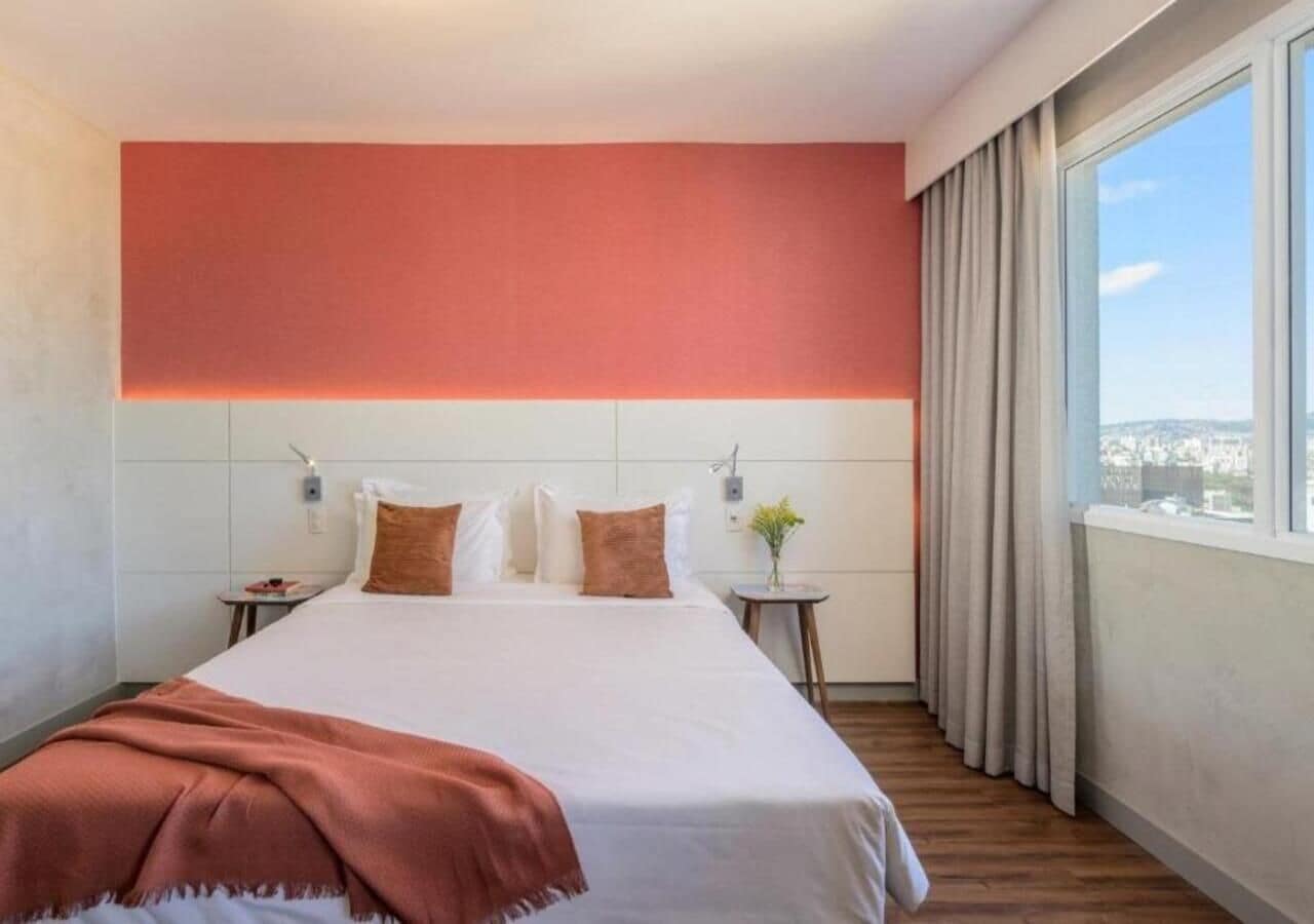 Un cuarto de hotel con una cama grande, una mesita de noche, una ventana con cortinas y vista a la ciudad.