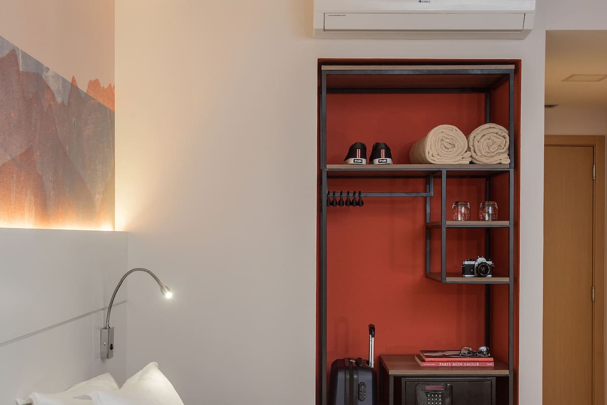 A hotel room with a bed, a desk with a lamp, a shelf with toiletries, and an air conditioning unit.