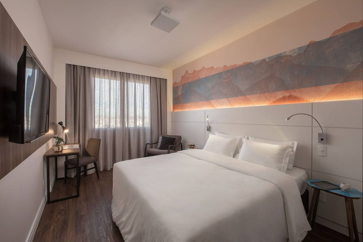 A hotel room with a large bed, a desk with a chair, a television, and a wall-mounted lamp.