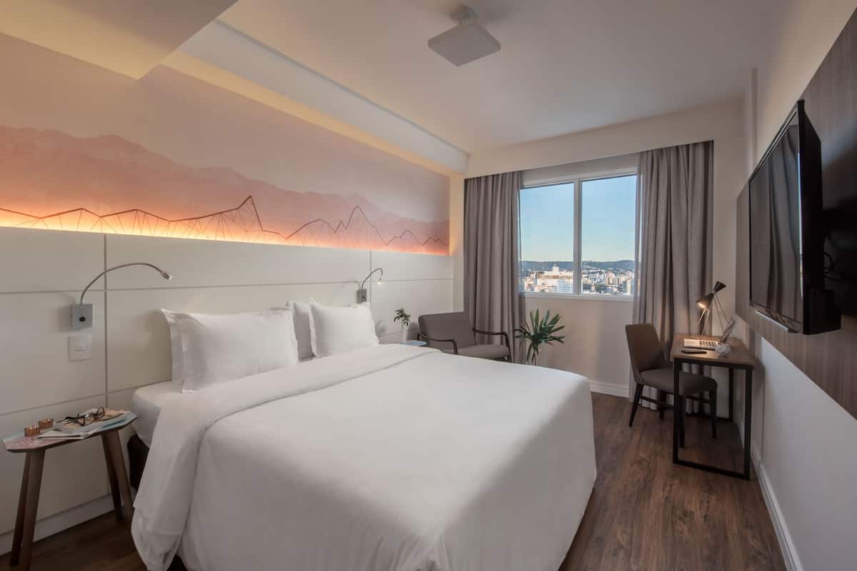 A modern hotel room with a large bed, a desk, a chair, a TV, and a city view through the window.