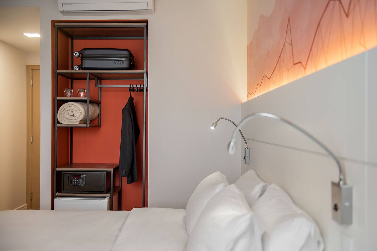 A hotel room with a bed, a wardrobe with shelves, a desk, and a wall-mounted lamp.