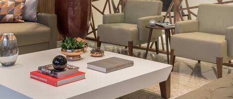 Un área de lounge moderna con una mesa de centro blanca, varios sillones, una planta grande en maceta y un diseño geométrico en la pared.