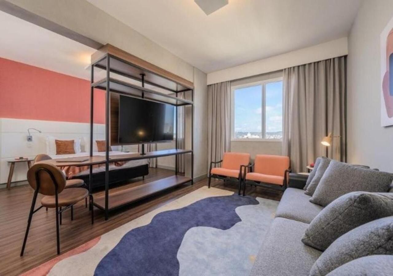 A modern hotel room with a flat-screen TV, a sofa, two chairs, a dining table, and a large window with curtains.
