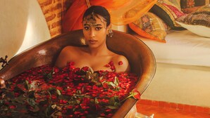 A person in a large copper tub filled with rose petals, set against a backdrop of a brick wall and a bed with colorful bedding. - Riad Persephone (Marrakech)