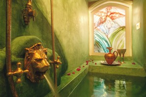 A bathroom with a green wall, a lion head faucet, a window with a flower painting, and a bathtub with rose petals.