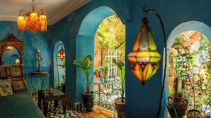 A room with a blue wall, a chandelier, a mirror, a table with a teapot, and potted plants. - Riad Persephone (Marrakech)