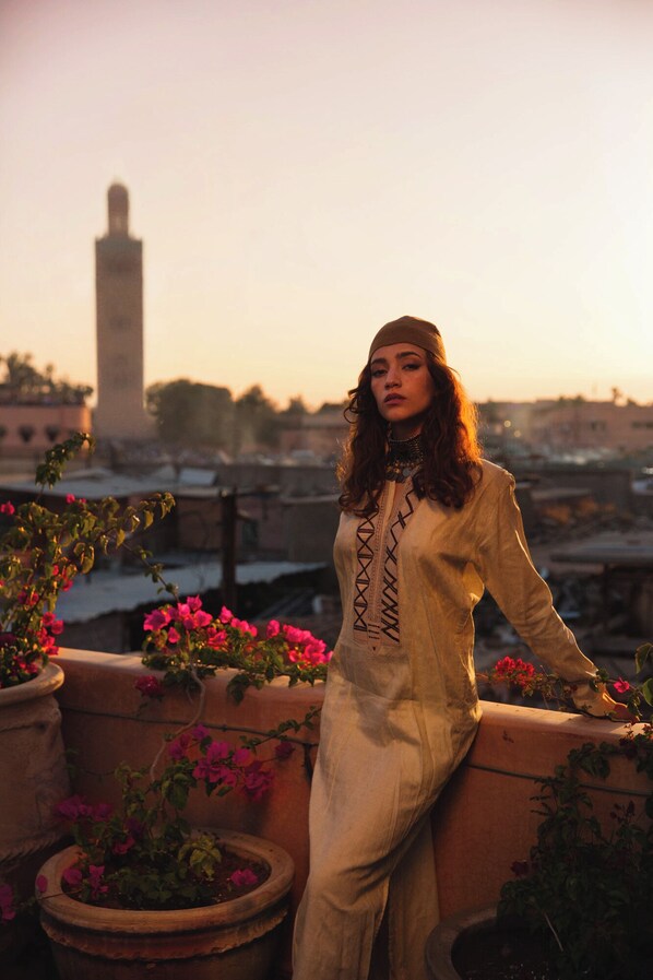 A person standing on a balcony with potted plants and flowers, overlooking a cityscape with a prominent tower in the background.