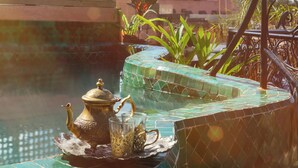 A rooftop terrace with a decorative teapot and glasses on a tiled table, surrounded by lush plants and a view of a distant tower. - Riad Persephone (Marrakech)