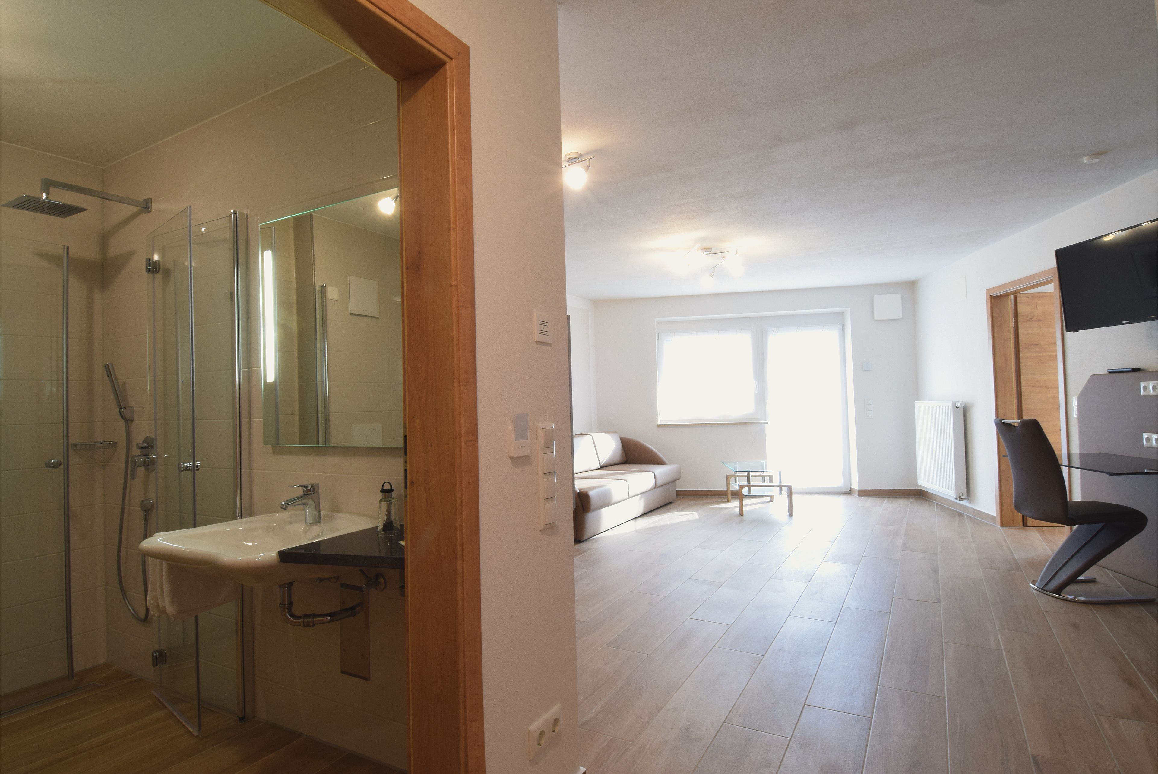 Un bagno moderno con box doccia in vetro, un lavandino e uno specchio. La stanza ha pavimento in legno e un'ampia finestra che garantisce luce naturale.
