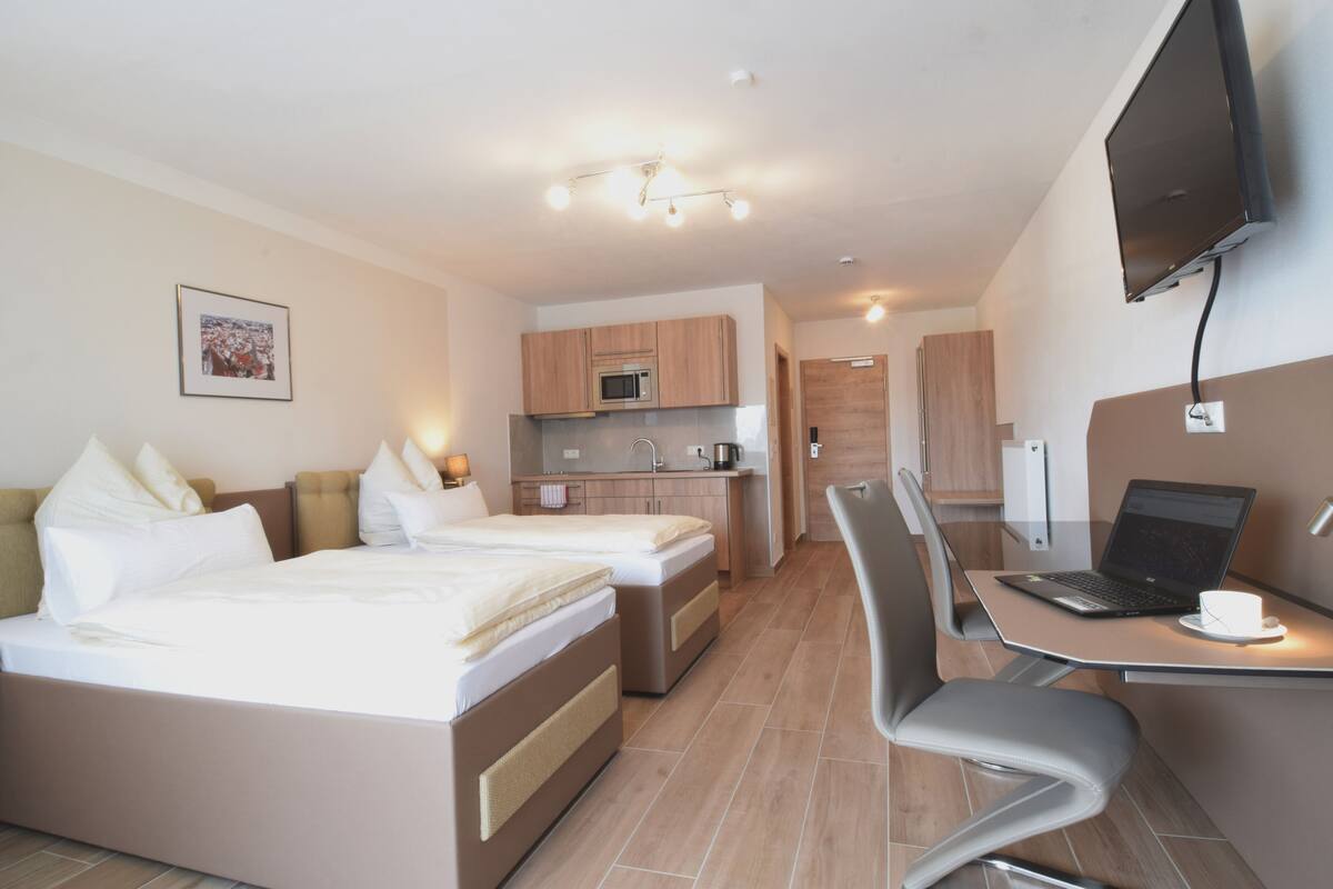 A modern hotel room with two beds, a kitchenette, a dining area with a laptop, and a TV mounted on the wall.