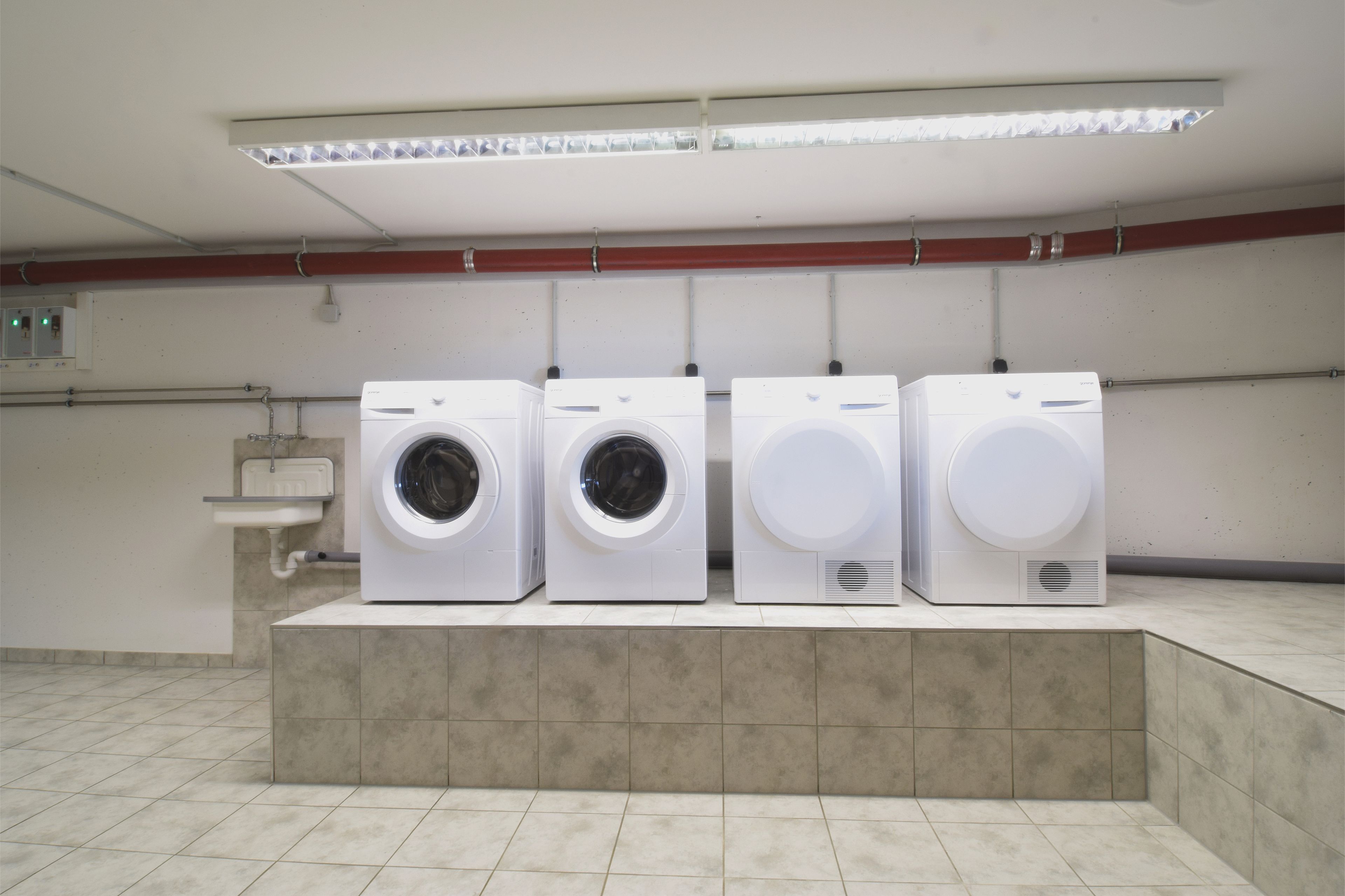 Een rij van vier witte wasmachines in een schone, betegelde ruimte met een wasbak en een rode leiding aan de muur.