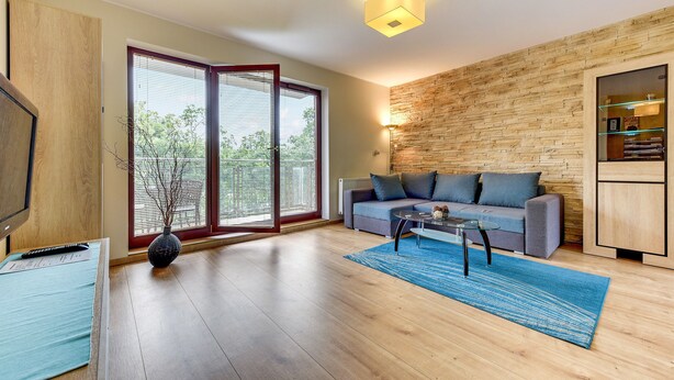 A modern living room with a sofa, coffee table, and a large window with blinds. - Rent a Flat Apartments - Torunska 18 (Gdansk)