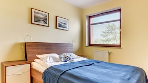 A bedroom with a wooden bed, a nightstand, a drawer, two framed pictures on the wall, and a window with blinds. - Rent a Flat Apartments - Torunska 18 (Gdansk)
