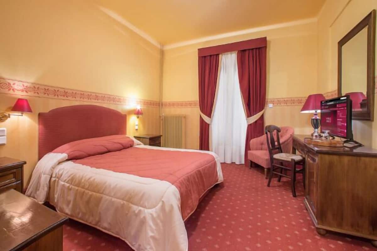 A hotel room with a large bed, a desk with a computer, a chair, and a red carpet.