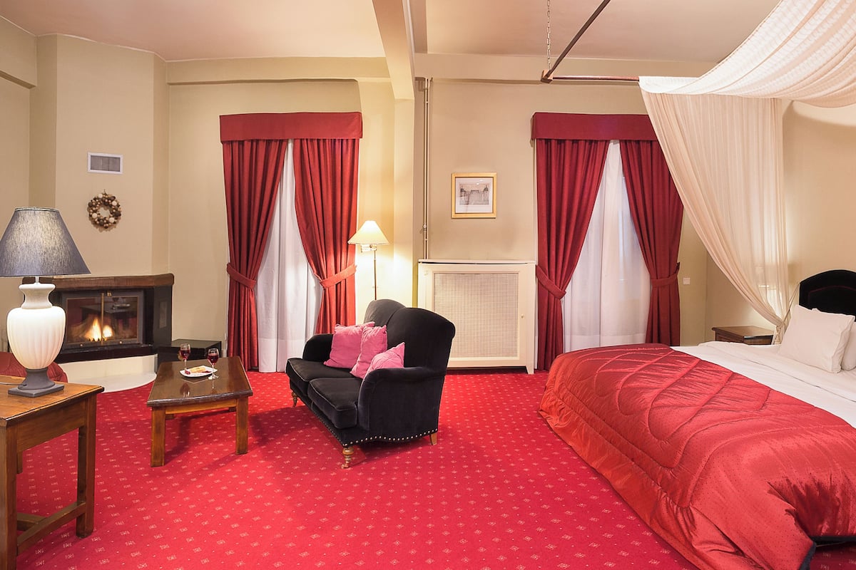 A hotel room with a bed, a chair, a table, a lamp, a fireplace, and curtains.