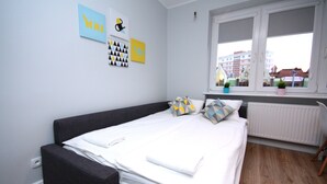 Une chambre moderne avec un lit, un canapé gris et une fenêtre donnant sur des immeubles.