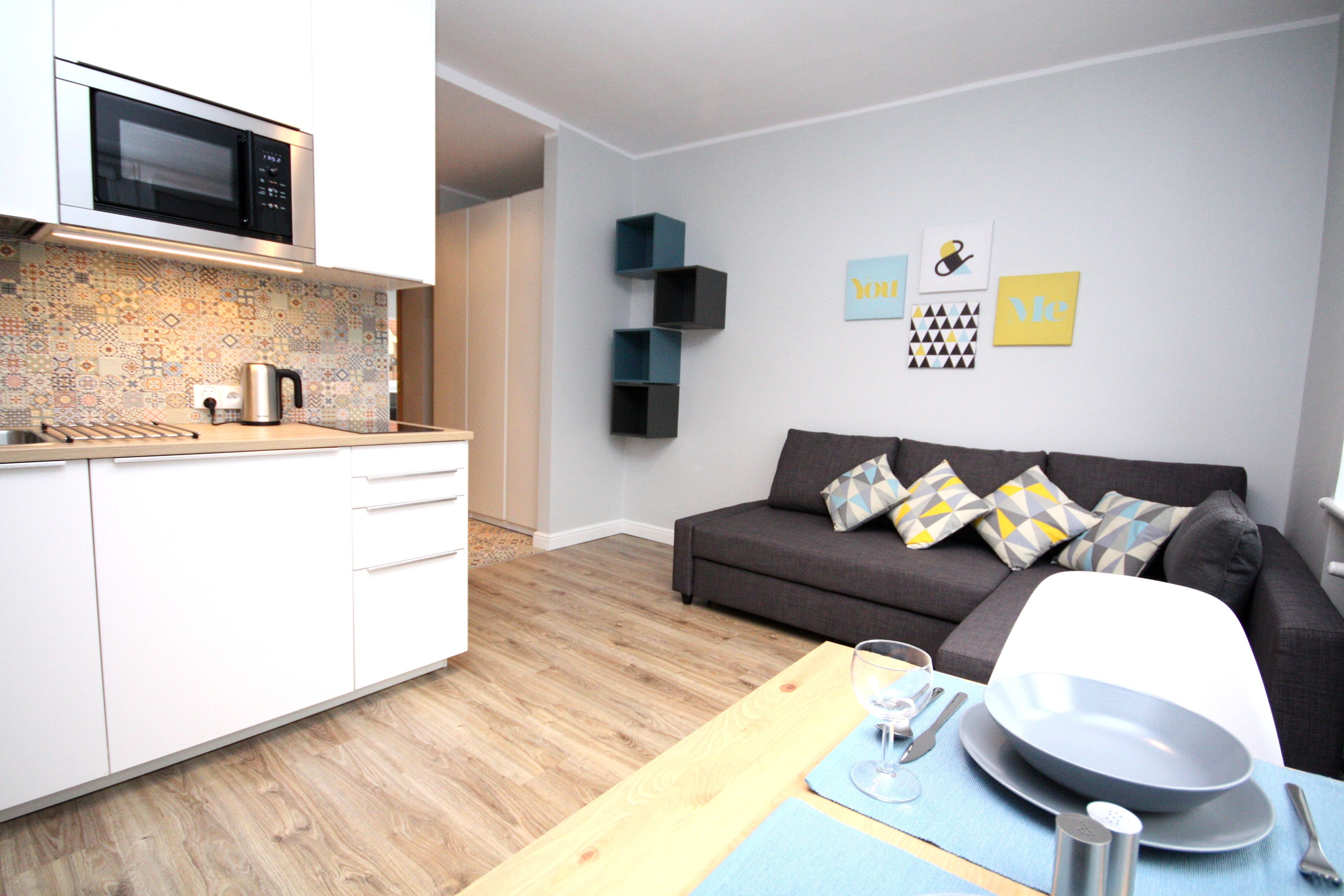 A modern kitchen with a microwave, a coffee maker, and a sink. There is a dining area with a table set for two, a sofa, and a wall with decorative items.