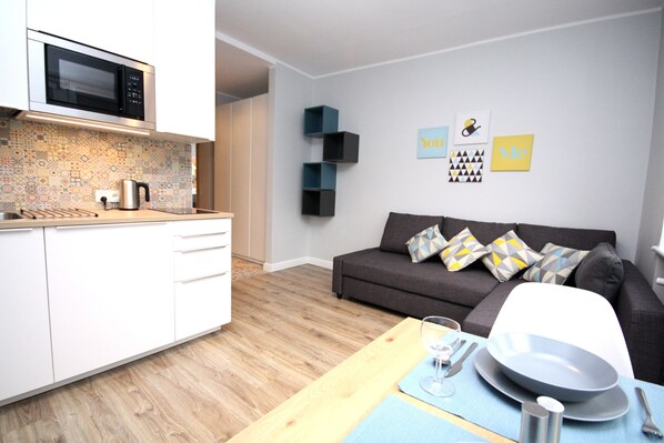 Una cocina moderna con microondas, cafetera y fregadero. Hay una zona de comedor con una mesa para dos personas, un sofá y una pared con objetos decorativos.
