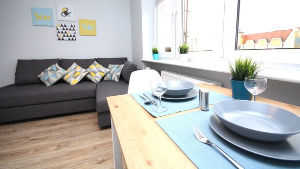 Un salon moderne avec un canapé gris, des coussins bleus, une table en bois dressée pour le repas et une fenêtre donnant sur des immeubles.