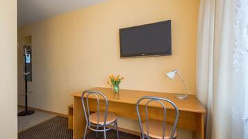 A hotel room with a wooden desk, two metal chairs, a TV mounted on the wall, a desk lamp, and a vase with flowers.