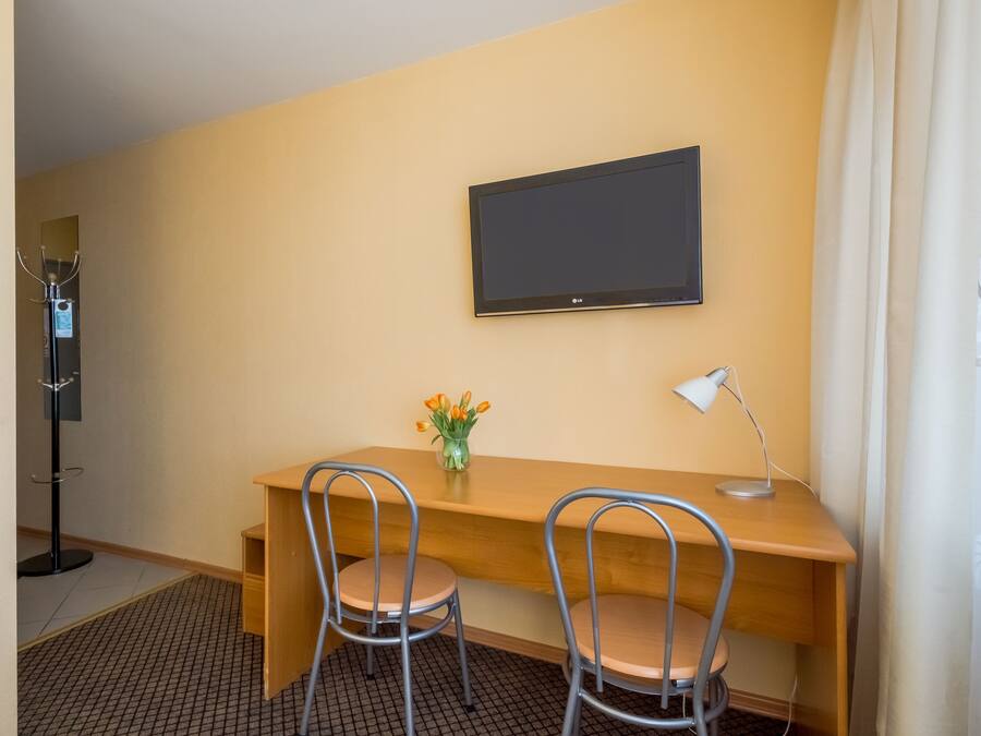 A hotel room with a wooden desk, two metal chairs, a TV mounted on the wall, a desk lamp, and a vase with flowers.