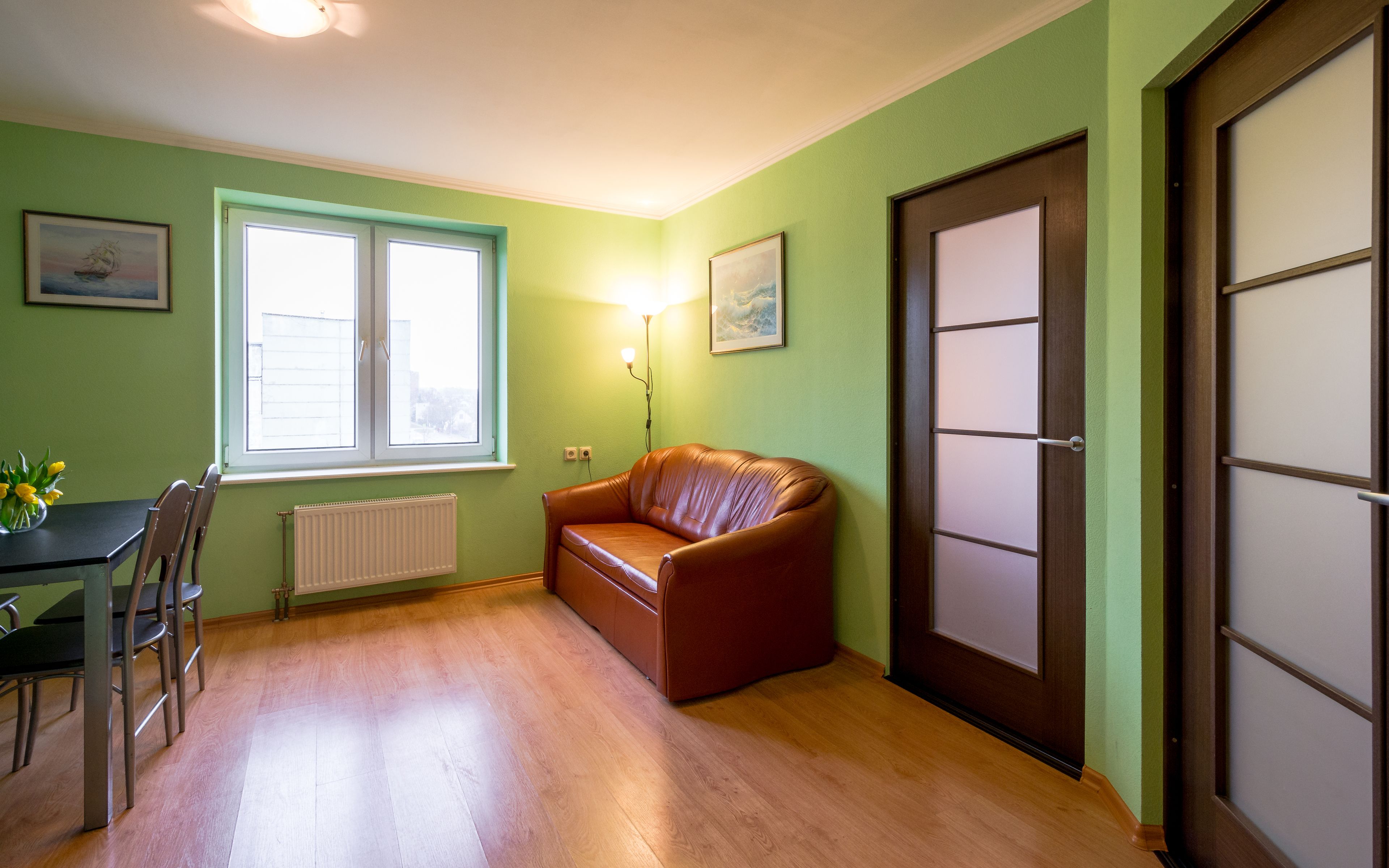 A room with a brown leather sofa, a dining table with chairs, a window with blinds, and a door with a frosted glass panel.