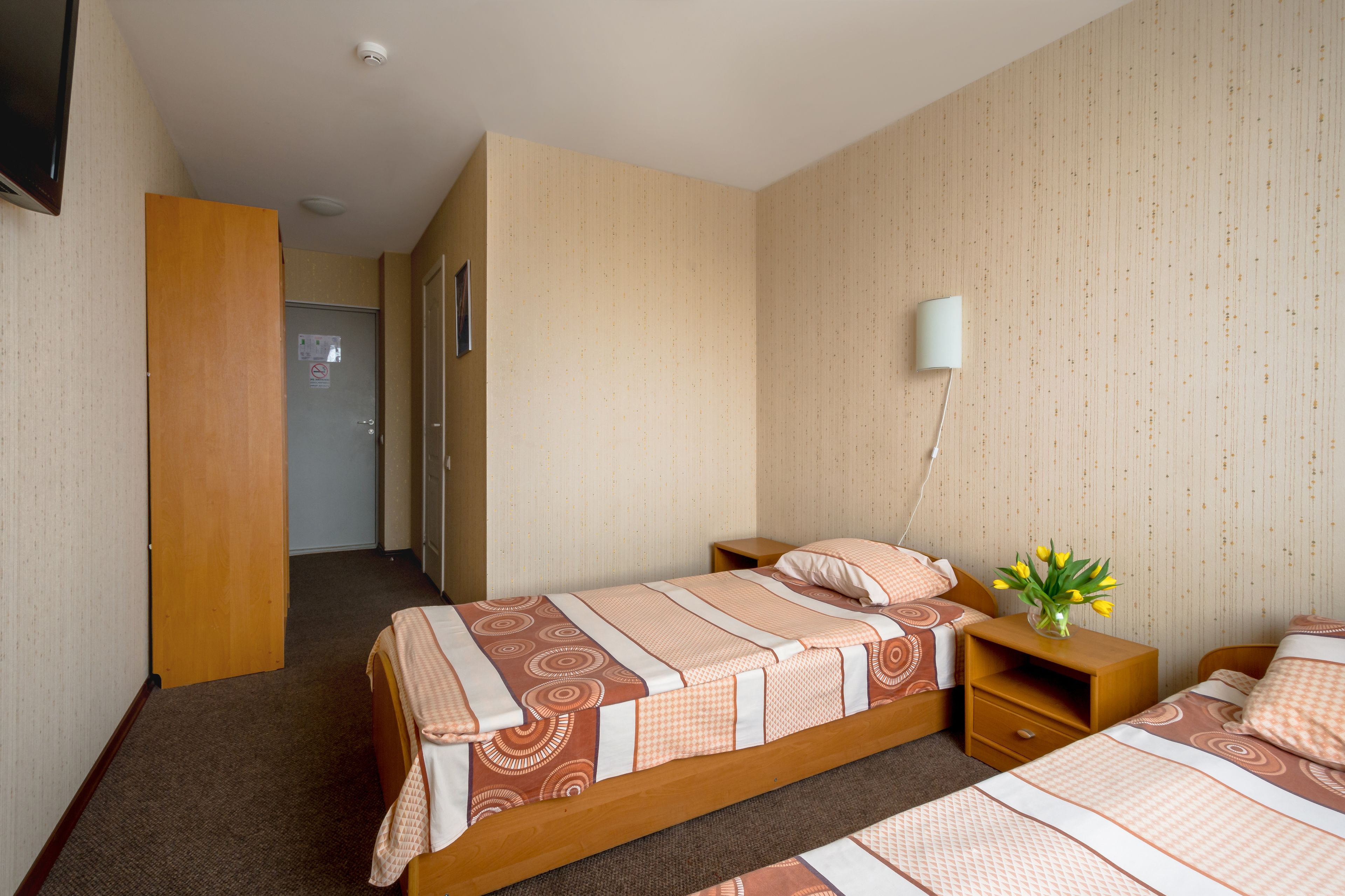 A hotel room with two beds, a wooden wardrobe, a bedside table with a vase of flowers, and a wall-mounted lamp.