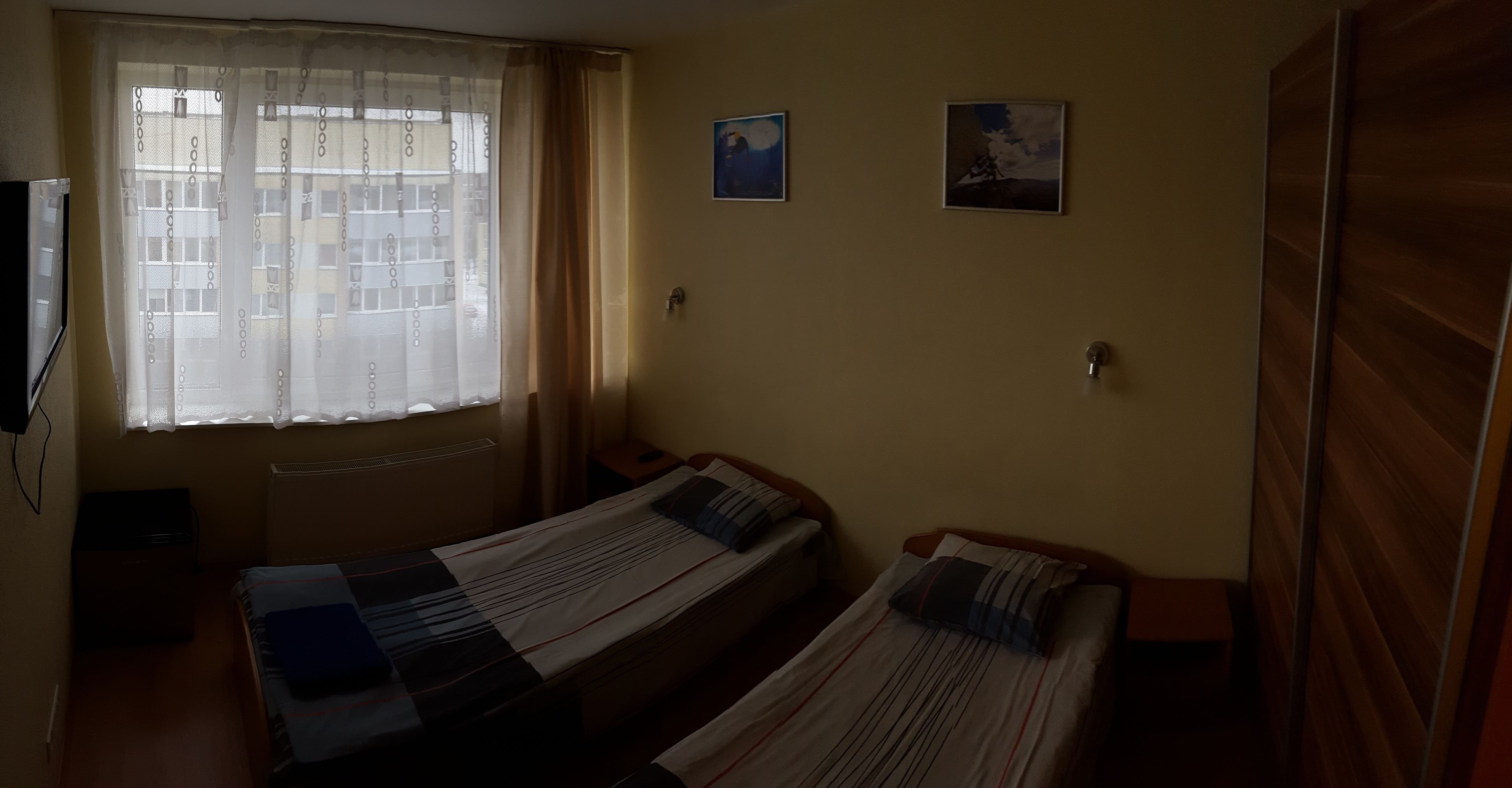 A room with two single beds, a window with sheer curtains, and two small wall-mounted pictures.