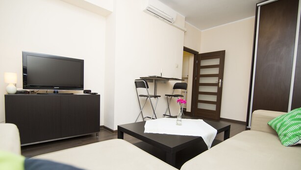 A modern living room with a flat-screen TV on a dark cabinet, a small table with a vase, two chairs, a sofa, and a wooden door. - Rent a Flat apartments - Korzenna St. (Gdansk)