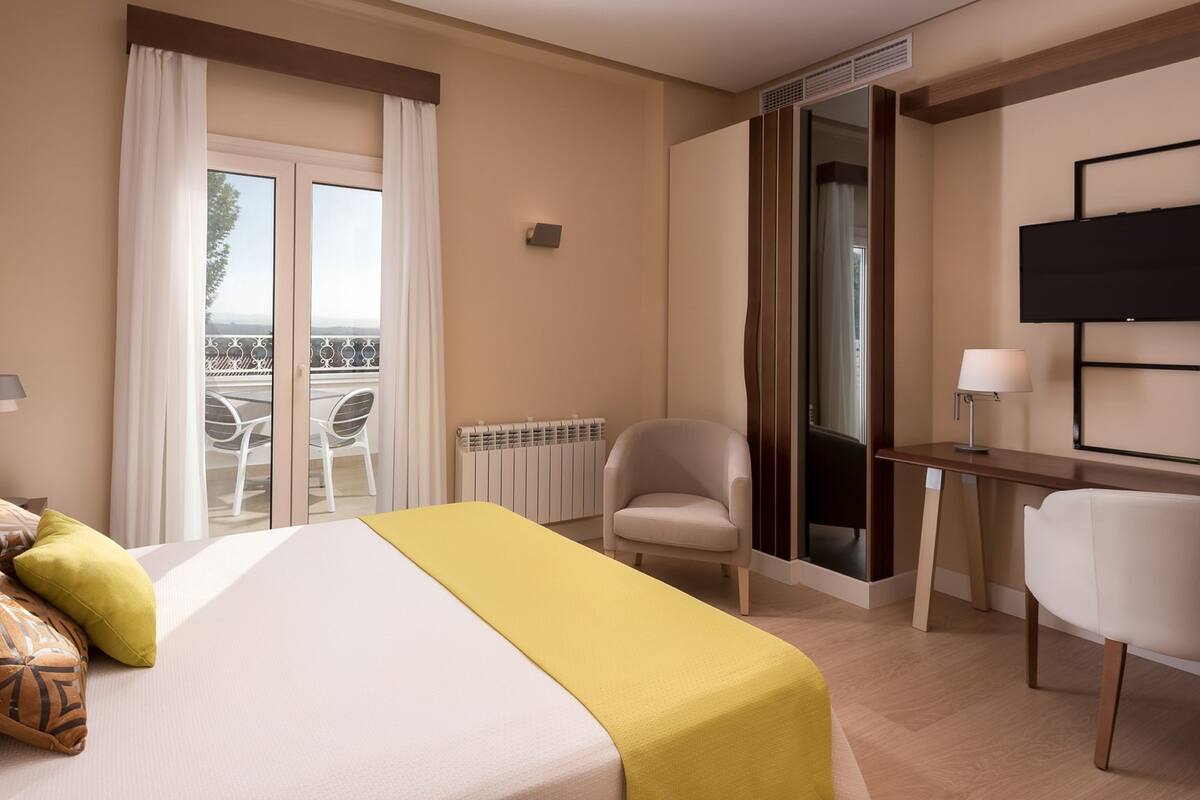 A hotel room with a bed, a desk, a chair, a TV, and a balcony with outdoor furniture.