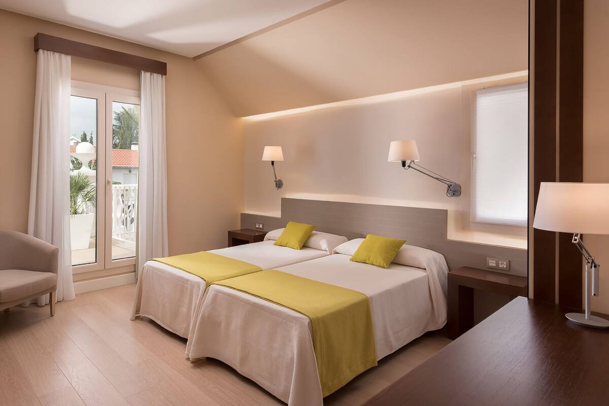 A hotel room with two beds, a seating area with chairs, a balcony with curtains, and two wall-mounted lamps.