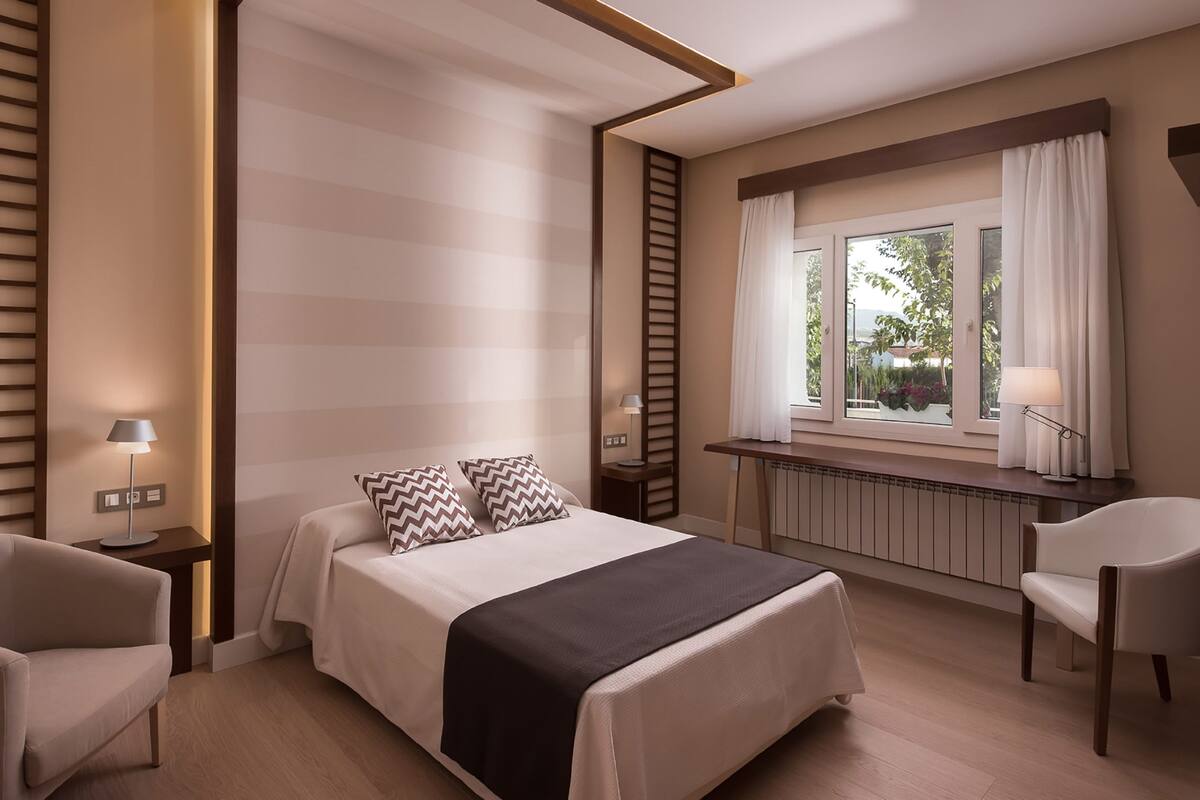 A modern hotel room with a bed, two chairs, a desk, and a window with curtains.
