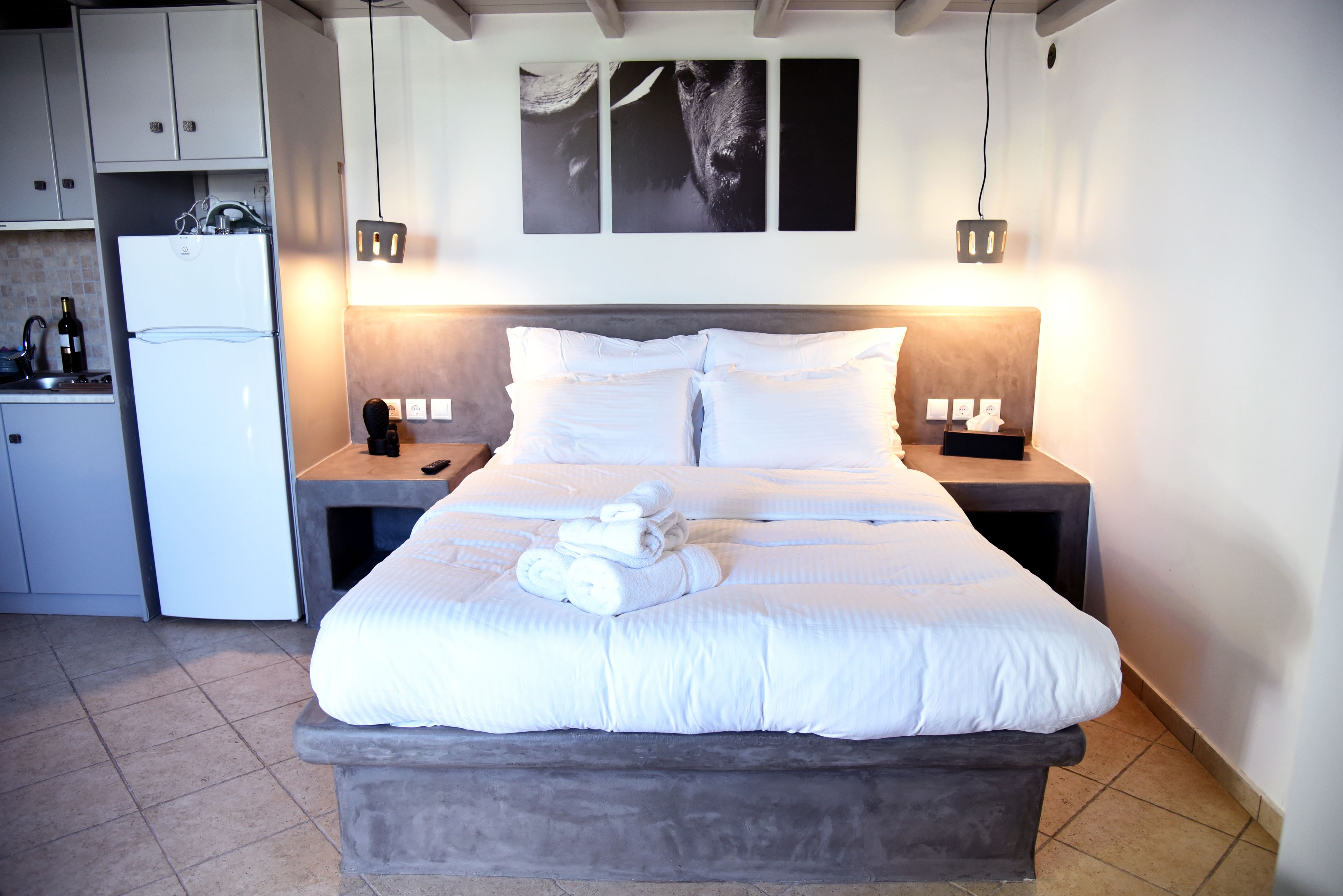 A bedroom with a bed, white bedding, a nightstand with a lamp, a small table with a bottle, and a refrigerator.