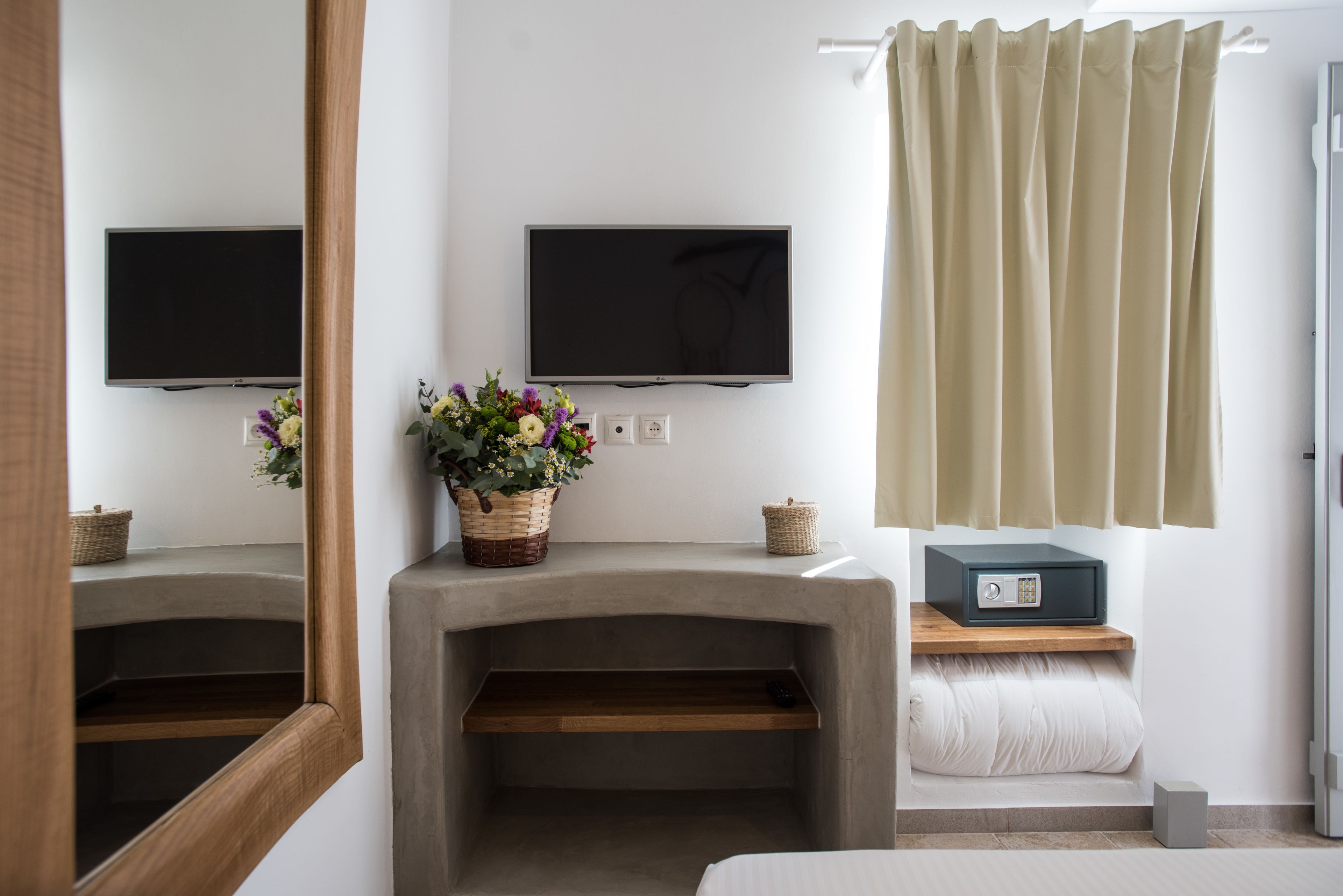 A hotel room with a television, a flower arrangement, a safe, and a window with curtains.