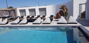 A pool area with lounge chairs and a hanging chair, surrounded by a white wall and a stone wall. - Aletro Cottage Houses (Mykonos)