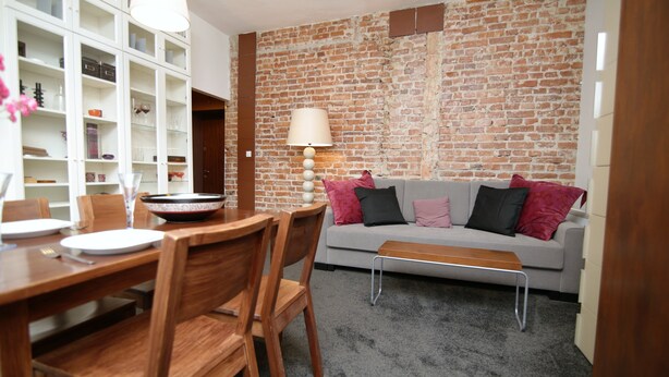 A dining area with a wooden table and chairs, a grey sofa with cushions, a lamp, and a brick wall. - Rent a Flat apartments - Ogarna St. (Gdansk)