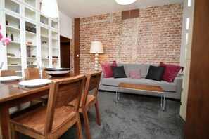 A dining area with a wooden table and chairs, a grey sofa with cushions, a lamp, and a brick wall. - Rent a Flat apartments - Ogarna St. (Gdansk)