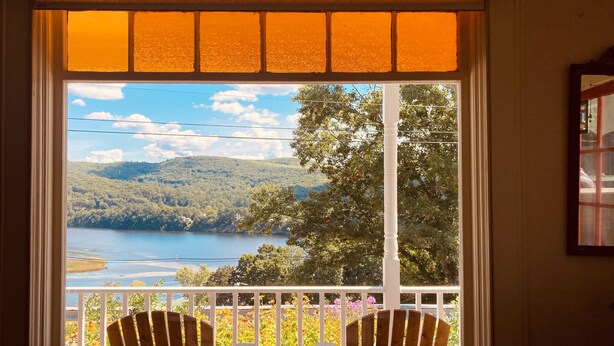 Une pièce dotée d’une grande fenêtre donnant sur un lac et des collines boisées. On y trouve deux chaises en bois sur un balcon.